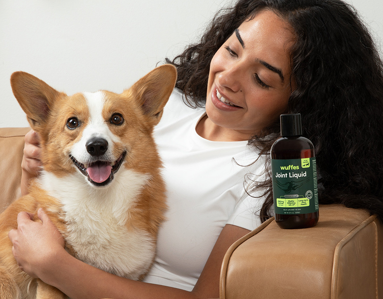 Woman with dark curly hair smiles while holding a happy corgi dog beside a bottle of joint supplement liquid
