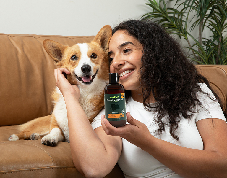 Woman holding omega-3 fish oil bottle next to smiling corgi on a leather couch